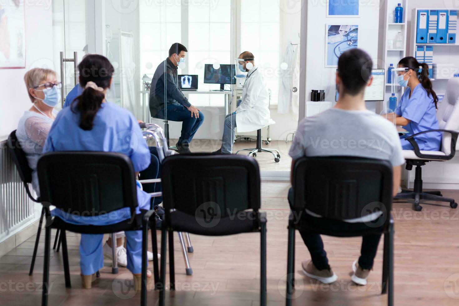 Doctor discussing with patient in hospital examination room wearing face mask against coronavirus and group of people in clinic lobby. Social distancing, medic, covid-19, man, worried, photo