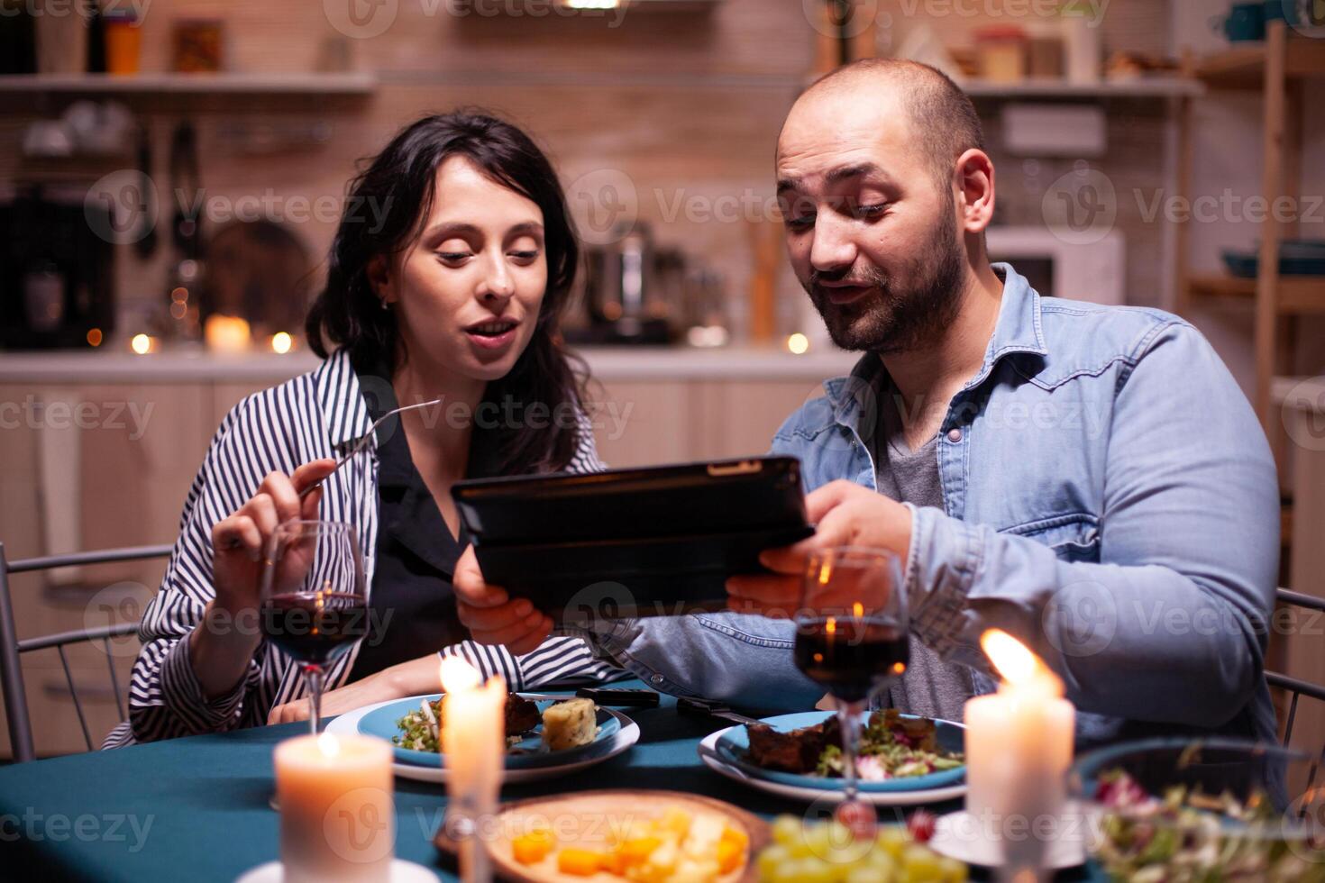 Cheerful young couple dining together using tablet during romantic dinner. Adults sitting at the table in the kitchen browsing, searching, , internet, celebrating, anniversary, happy., festive, enjoy. photo