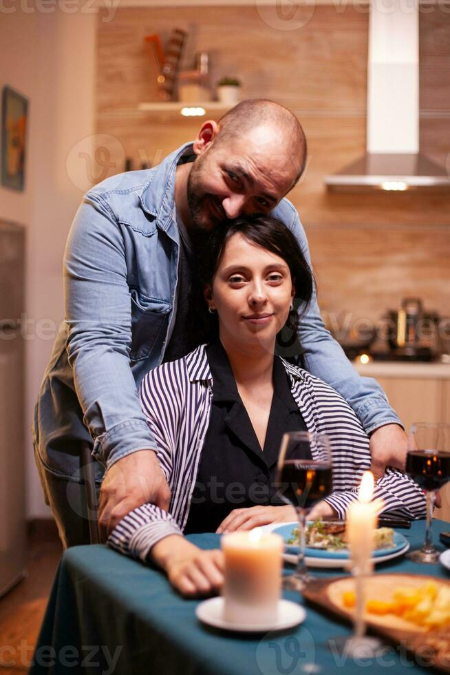 Couple celebrating relationship looking at webcam in kitchen. Married people special tender moments, enjoying the meal at candle lights celebration. photo