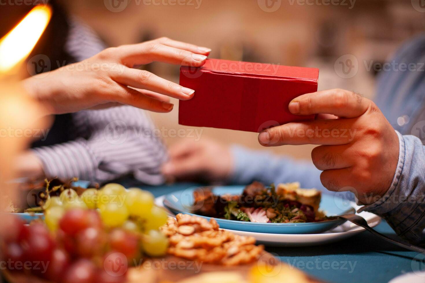 Man giving wife gift box and having romantic dinner together. Cheerful man dining with woman at home, enjoying the meal, celebrating their anniversary at candle lights. photo