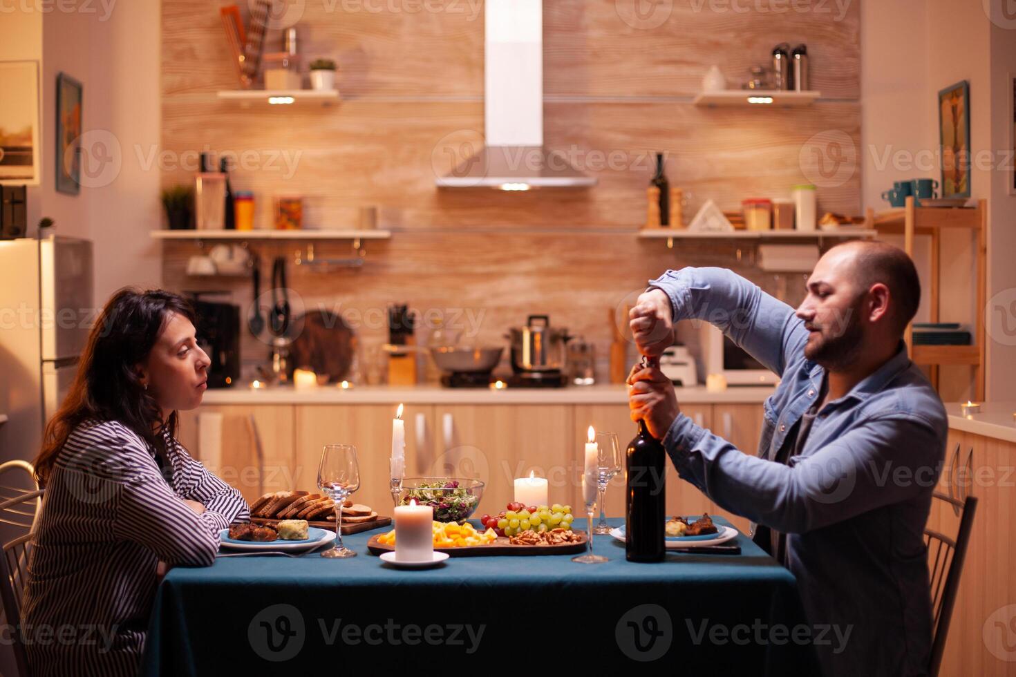Husband opens wine bottle while celebrating relationship with wife in kitchen. Happy couple talking, sitting at table in kitchen, enjoying the meal, celebrating their anniversary at home. photo