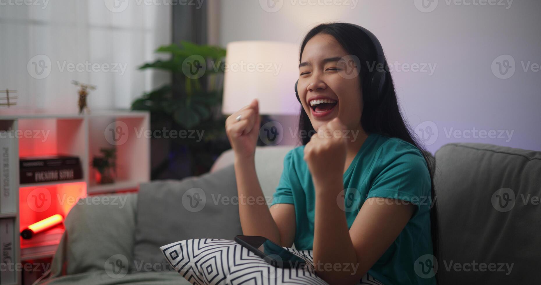 Portrait of Young Asian woman wearing glasses and headphones enjoys playing online esport games on smartphone sitting on sofa in the living room at home,Gamer lifestyle concept. photo