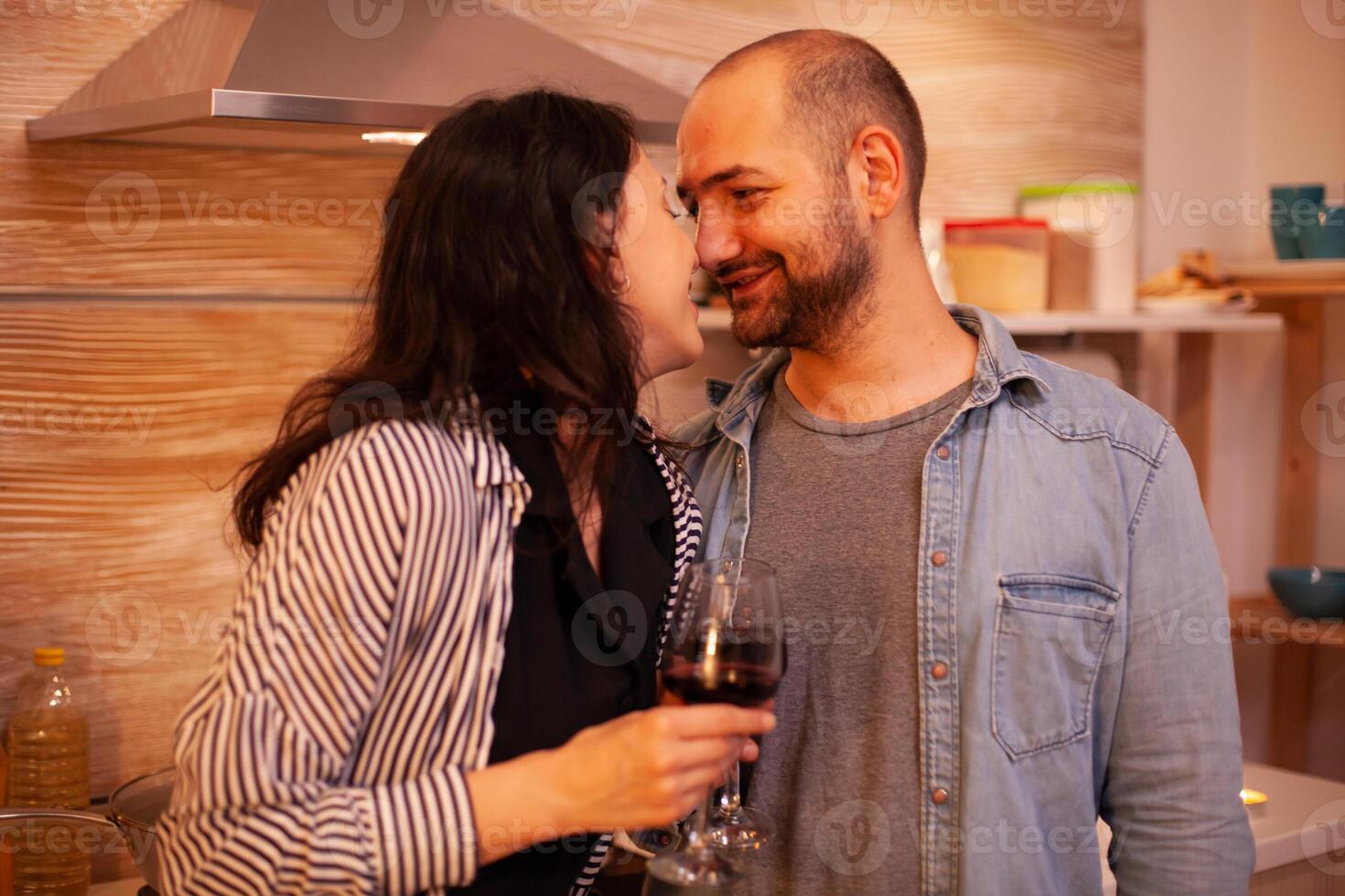 Couple touching noses and having romance date in kitchen. Adult couple at home, drinking red wine, talking, smiling, enjoying the meal in dining room. photo