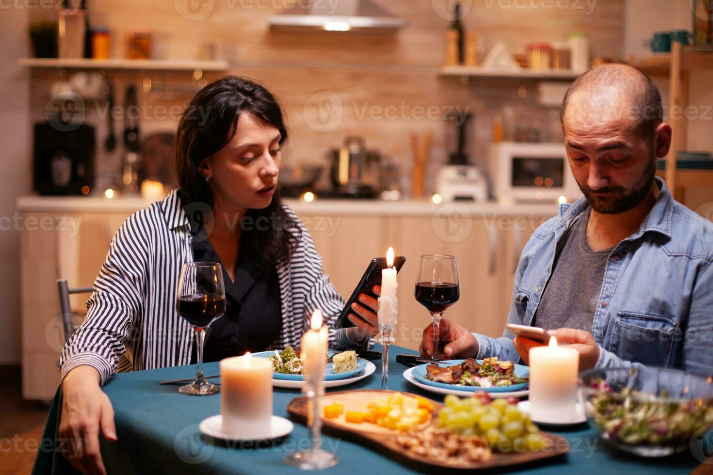 contento Pareja acecho vídeo en teléfono inteligente en cocina durante aniversario. adultos sentado a el mesa en el cocina hojeada, buscando, utilizando teléfonos inteligentes, Internet, celebrando aniversario. foto