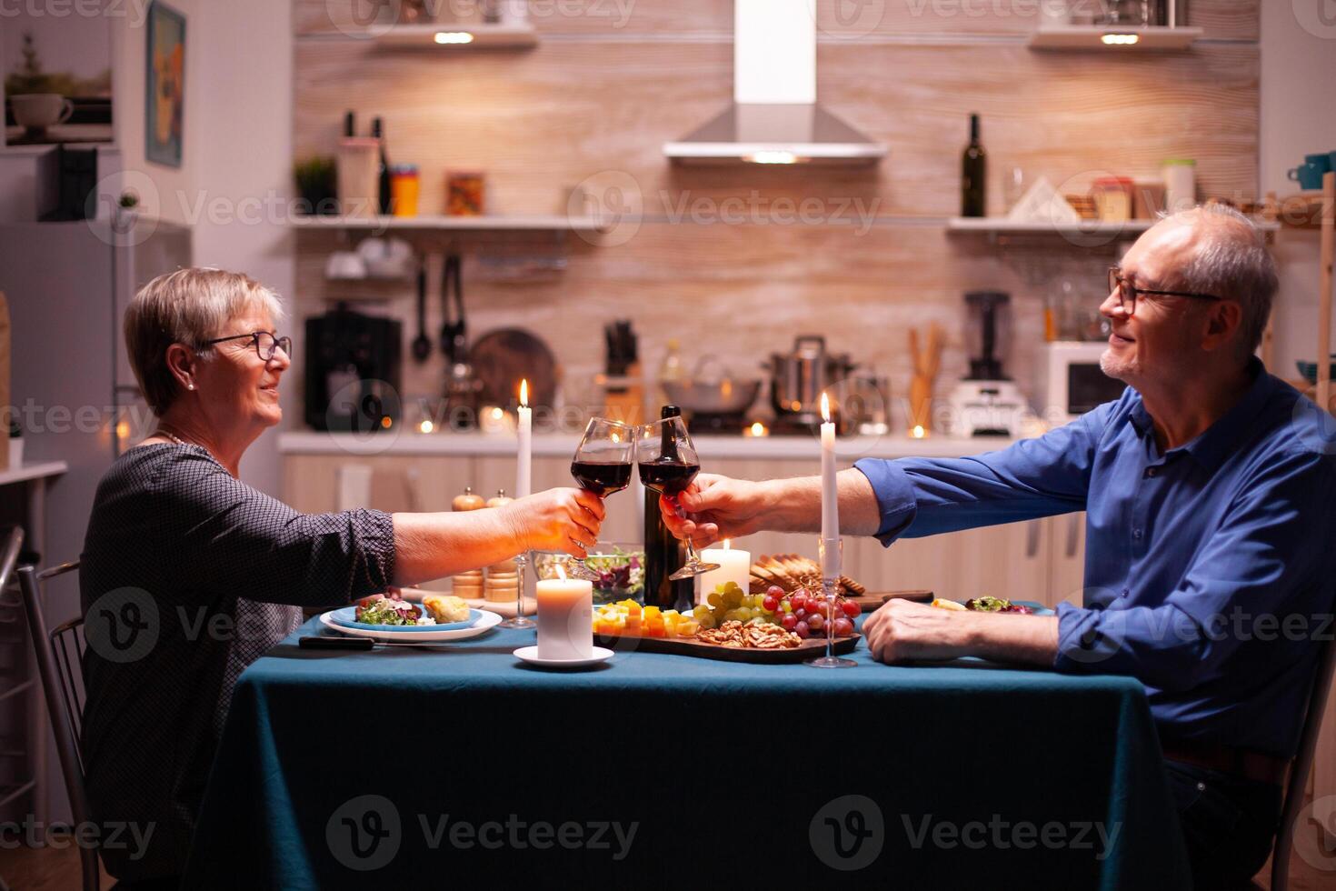 Senior couple toasting glasses with red wine during dinner. Elderly man and woman sitting at the table in kitchen, talking, enjoying the meal, celebrating their anniversary in the dining room. photo