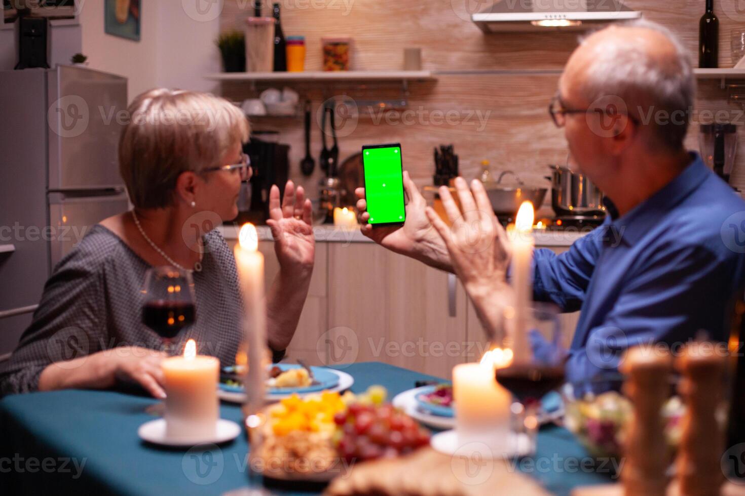 Elderly man and woman waving at phone with green screen. Aged people looking at mockup template chroma key isolated smart phone display using techology internet sitting at the table in kitchen. photo