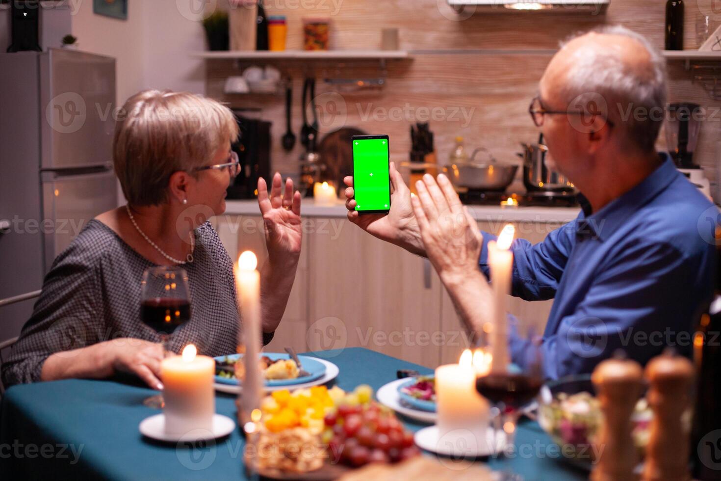 Aged man holding phone with green screen and wife is waving at it. Aged people looking at mockup template chroma key isolated smart phone display using techology internet sitting at the table photo