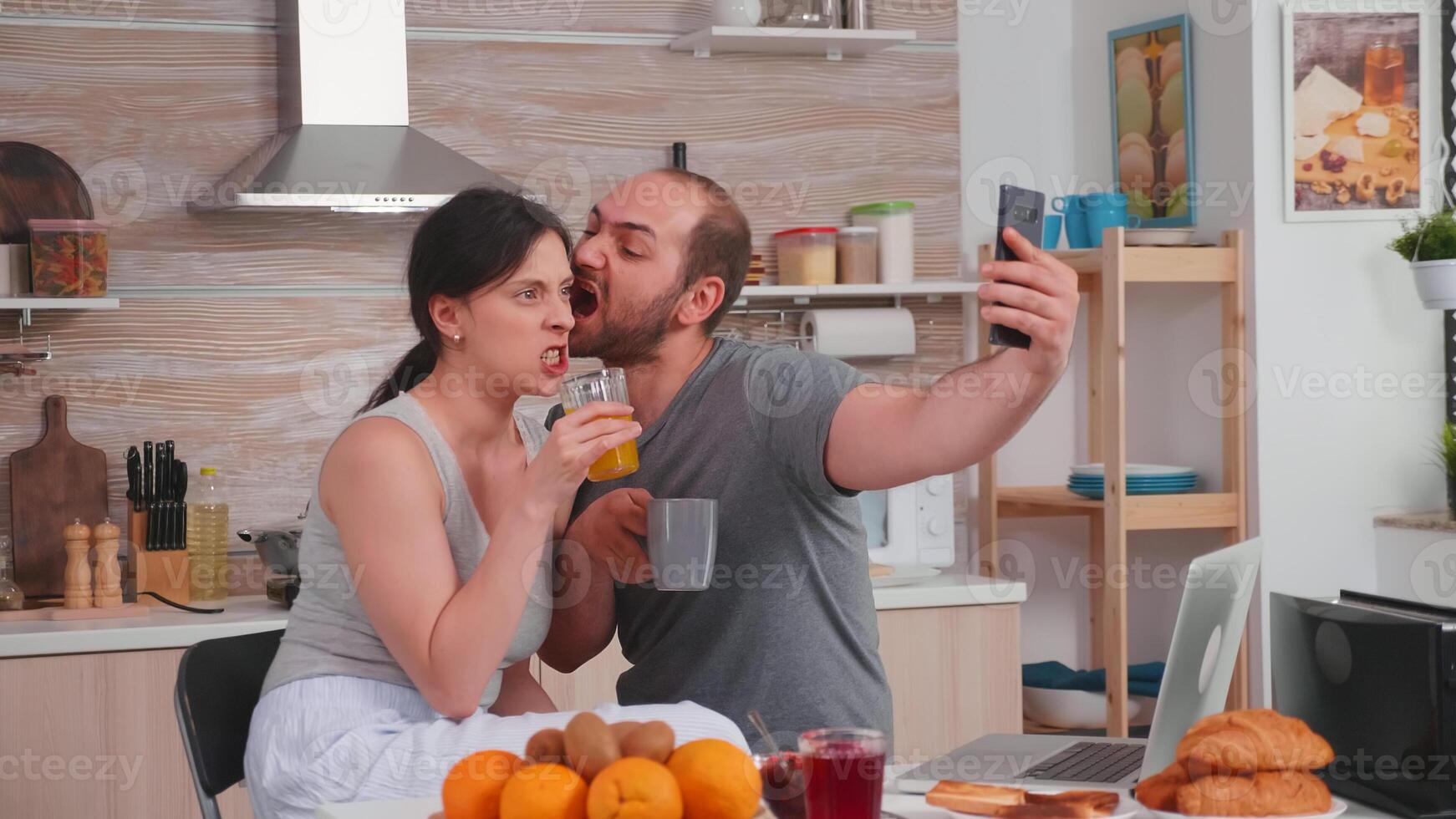 Happy young couple taking selfies during breakfast. Joyful married husband and wife making funny faces while taking a photo during breakfast in kitchen.