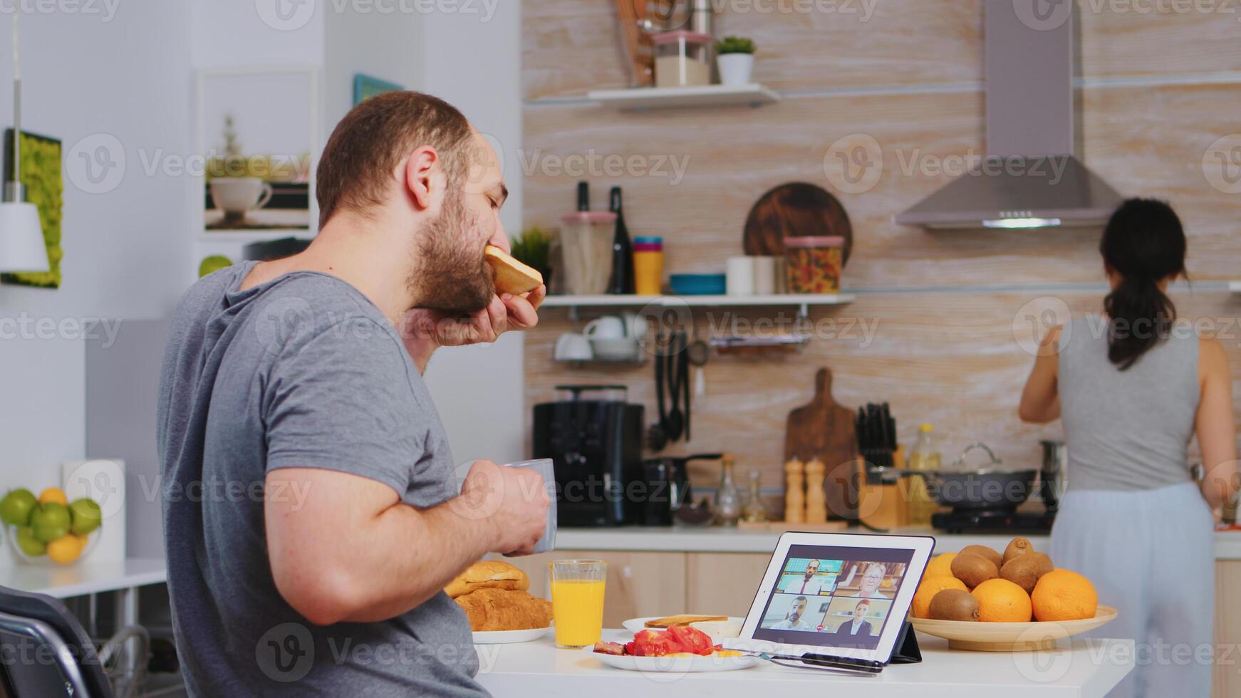 Entrepreneur on a video conference while eating breakfast in kitchen. Freelancer working remote, talking in videoconference video call online web internet meeting from home, communication device photo
