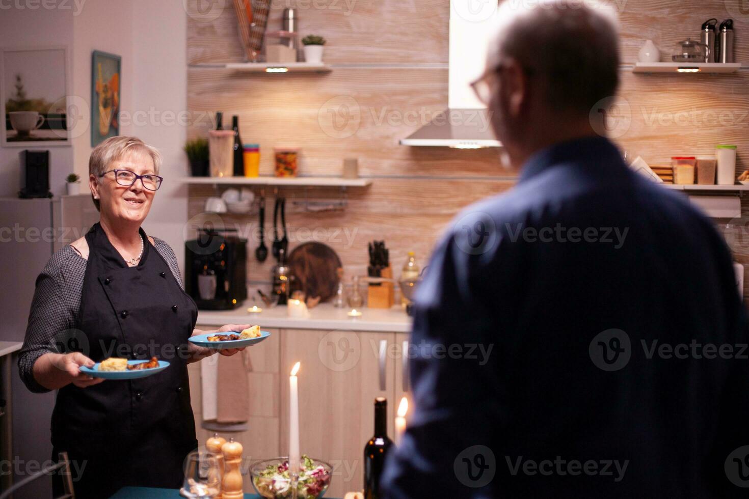 mayor mujer servicio marido con delicioso comida para su relación aniversario. mayor antiguo Pareja hablando, sentado a el mesa en cocina, disfrutando el comida, celebrando. foto