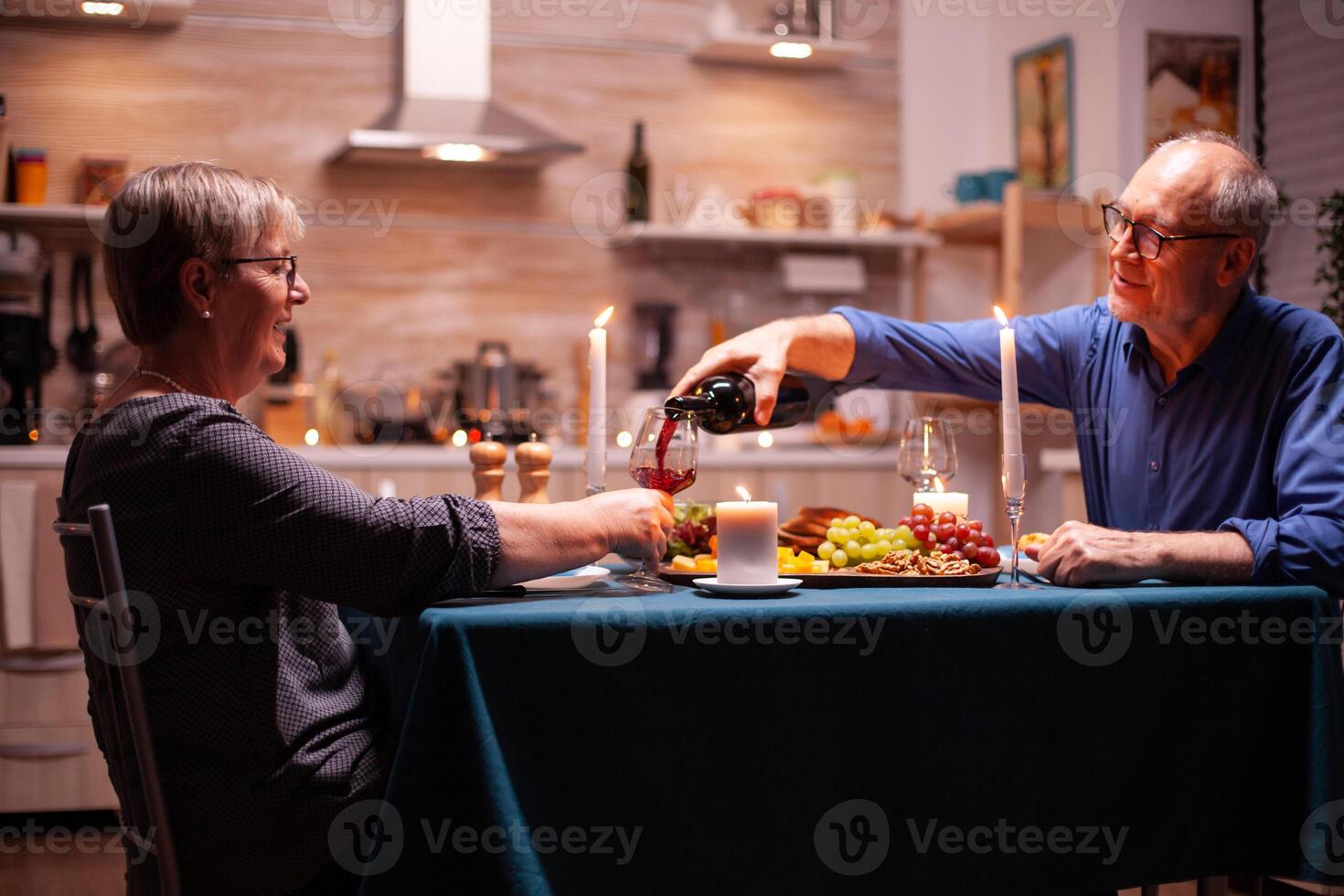 Senior man serving wife with wine during romantic dinner. Elderly couple sitting at the table in kitchen, talking, enjoying the meal, celebrating their anniversary in the dining room. photo
