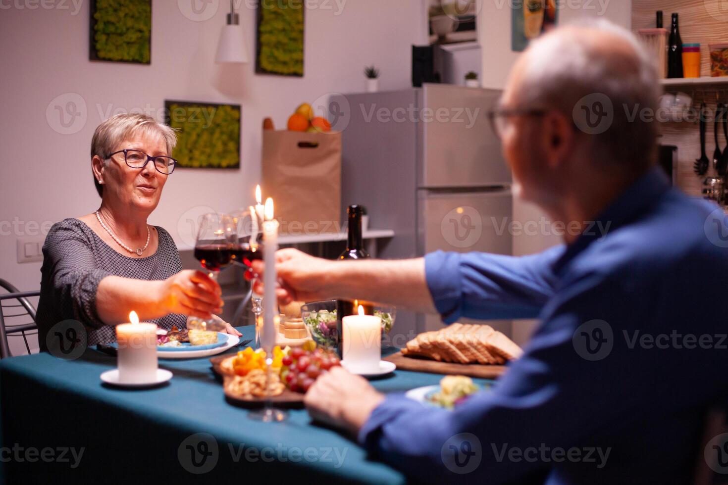 Senior couple clinking glasses with red wine during evening in kitchen. Elderly couple sitting at the table in kitchen, talking, enjoying the meal, celebrating their anniversary in the dining room. photo