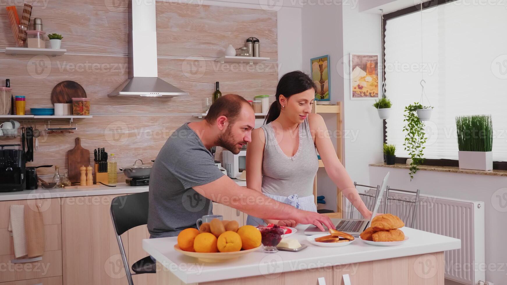 Young couple using laptop at the kitchen. Happy married husband and wife in pajamas using internet web online modern technology, smiling and happy in the morning. Reading news browsing internet photo