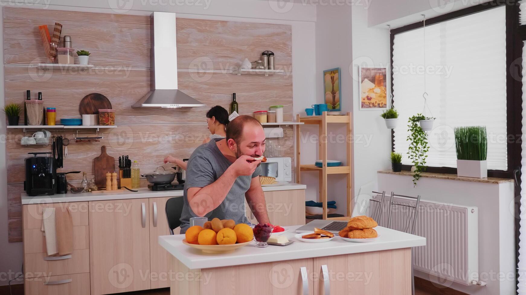 Freelancer drinking coffee and working on laptop during breakfast in kitchen. Entrepreneur in pajamas working on a laptop from home in kitchen. photo