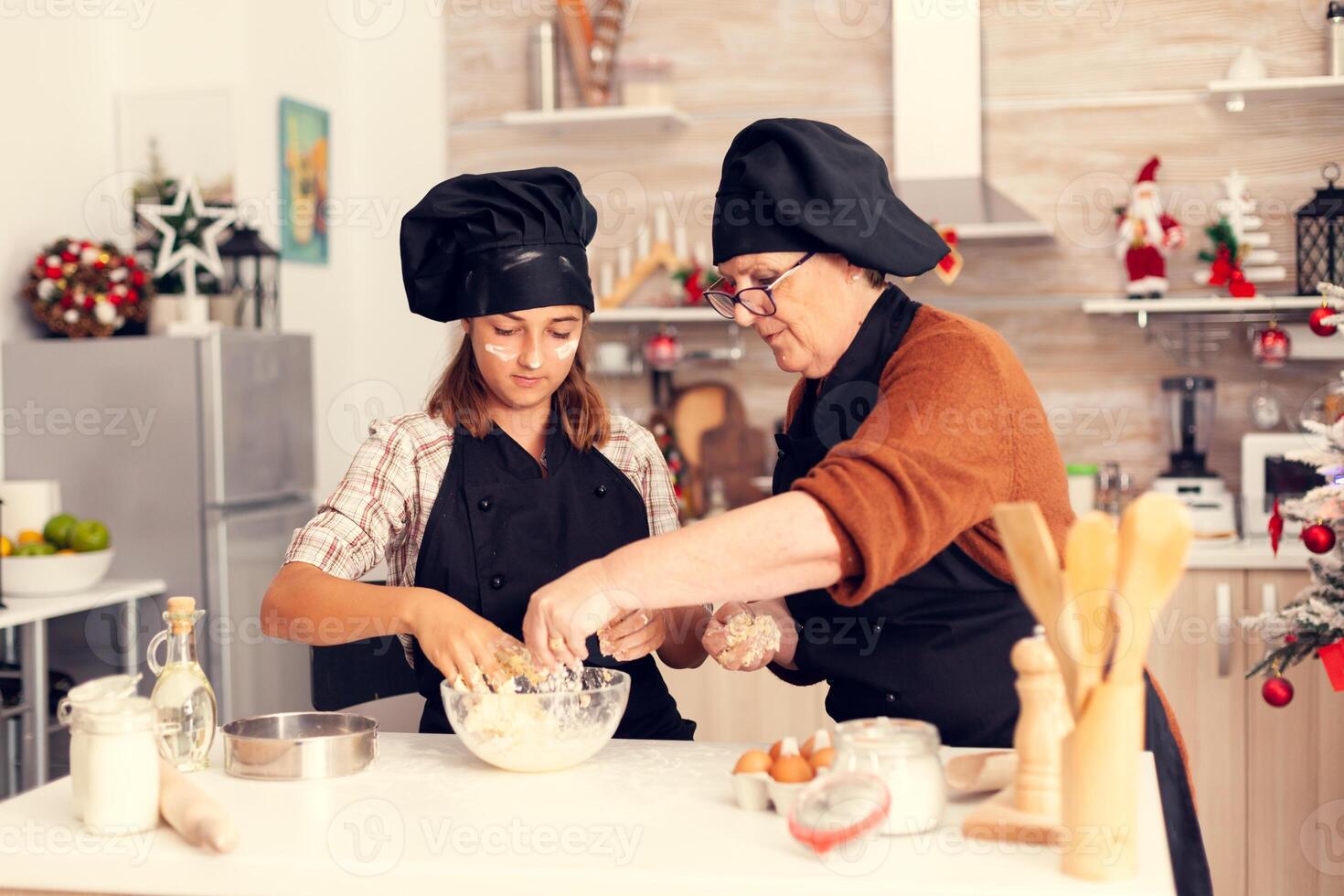 Kid making traditional cookies on christmas day wearing apron and bonette. Happy cheerful joyfull teenage girl helping senior woman preparing sweet biscuits to celebrate winter holidays. photo