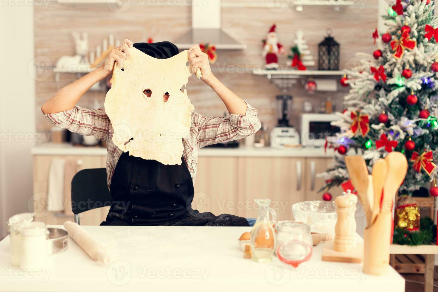 Cute kid doing traditional dessert on christmas day wearing apron and bonette. Cheerful happy cute girl while prepearing delicious cookies for christmas celebration in kitchen with christmas tree in the background. photo