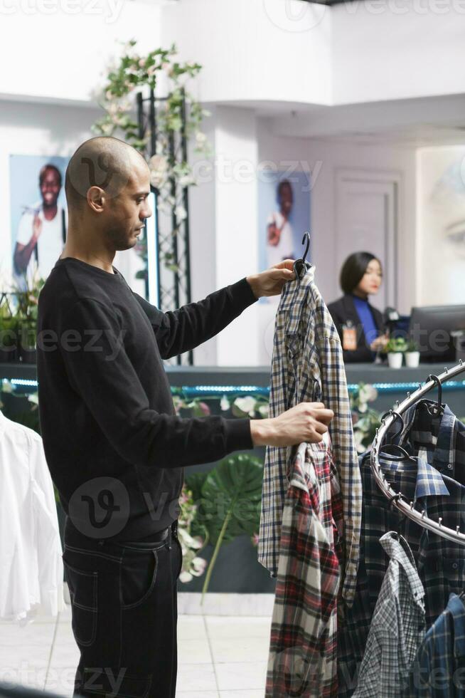 Young arab man holding two plaid shirts on hangers while shopping for casual outfit in shopping mall fashion department. Clothing store client examining garment style and size photo