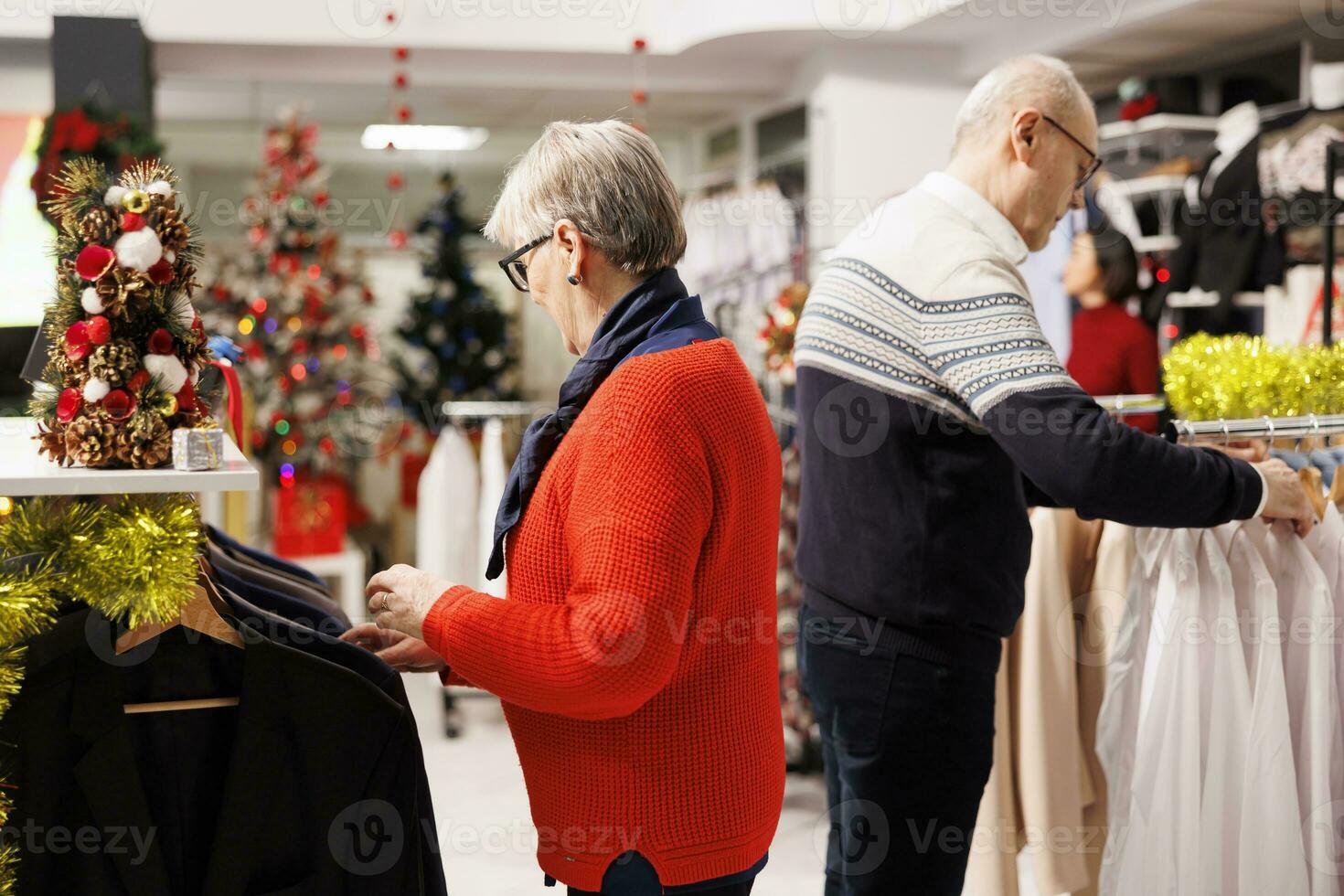 Elderly couple searching for blazers in clothing store with festive decorations, buying items as presents for family on christmas eve. Senior man and woman browsing through merchandise on discount. photo