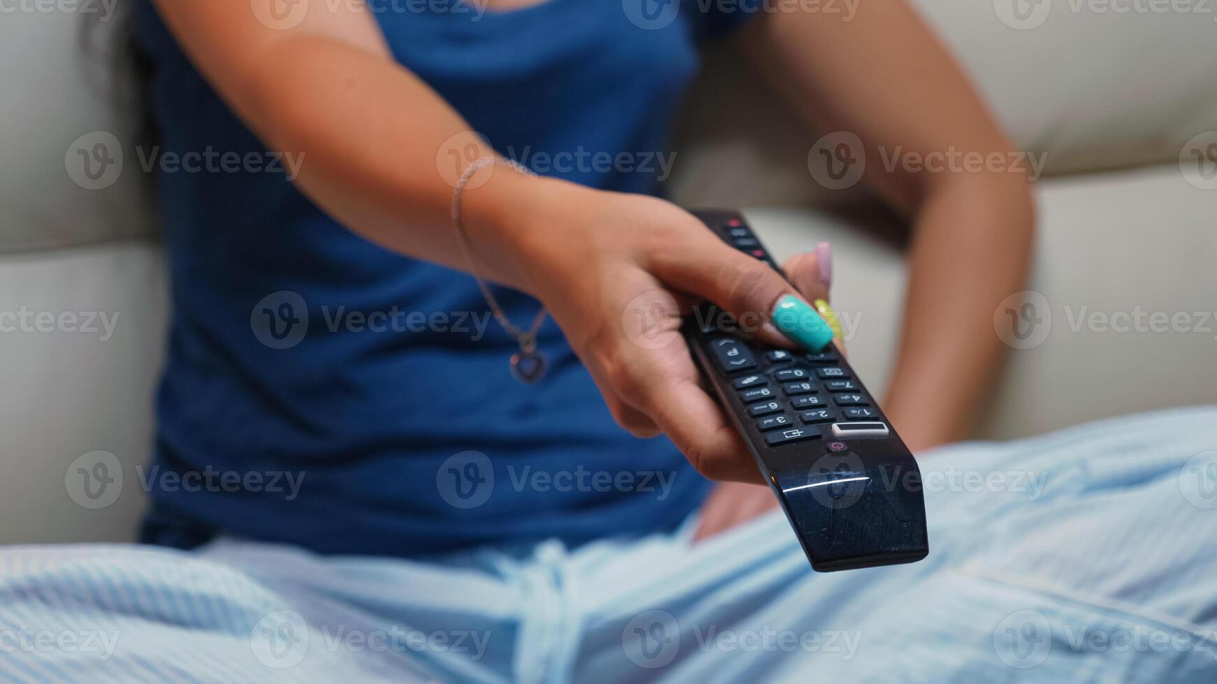 Close up of switching tv programs sitting on sofa using remote control. Shot of controller in hands of person pointing to TV pressing the button and changing channels sitting in front of television photo