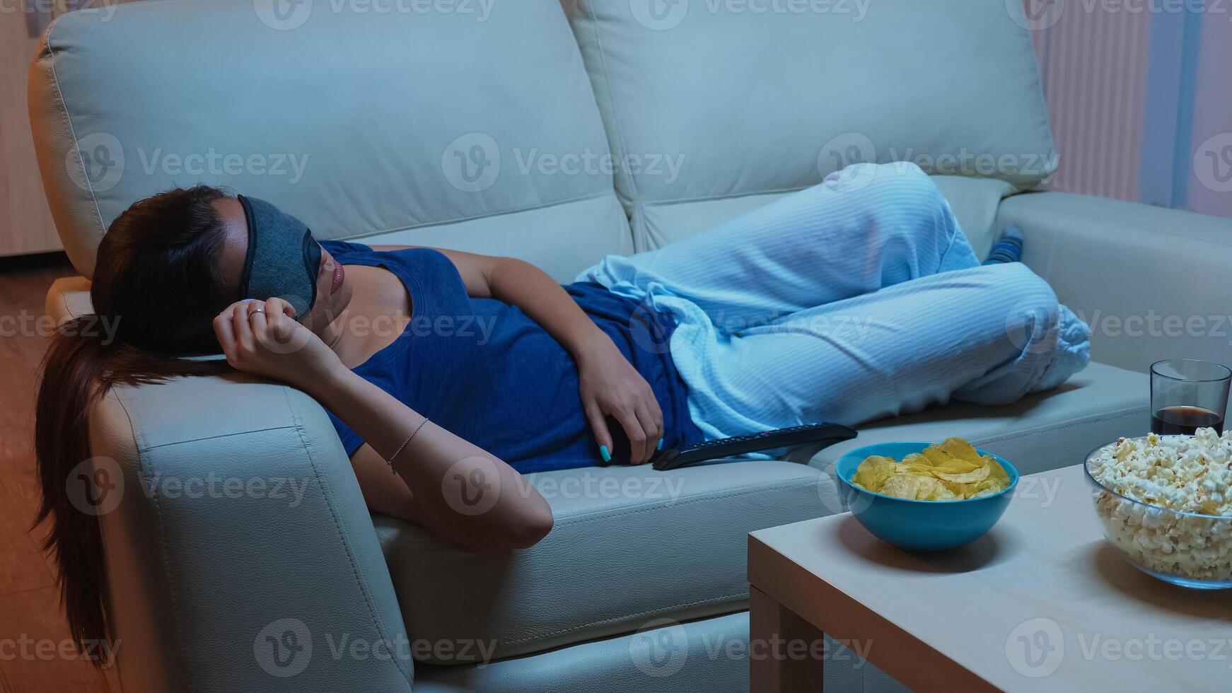 Sleeping on sofa with eye covering mask in front of television. Tired exhausted lonely sleepy woman in pajamas falling asleep on sofa during the movie, closing eyes while watching tv in living room. photo