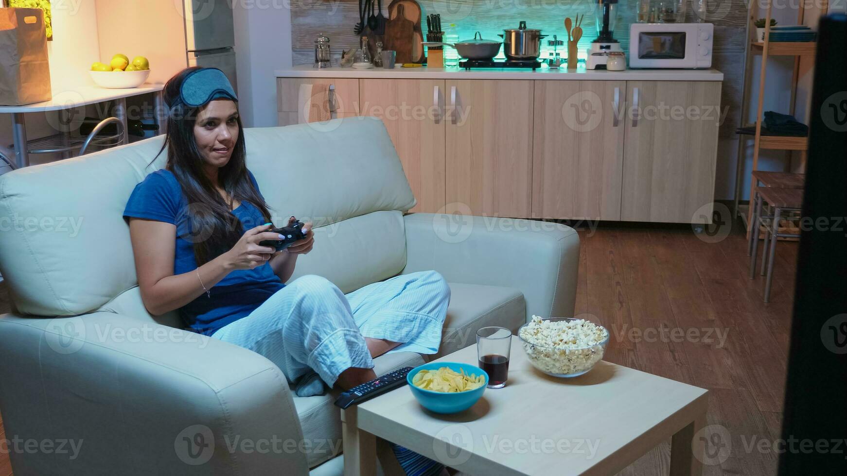 Woman sitting on sofa playing video game late at night wering eye mask on forehead. Excited determined gamer using controller joysticks keypad playstation gaming and having fun winning electronic game photo