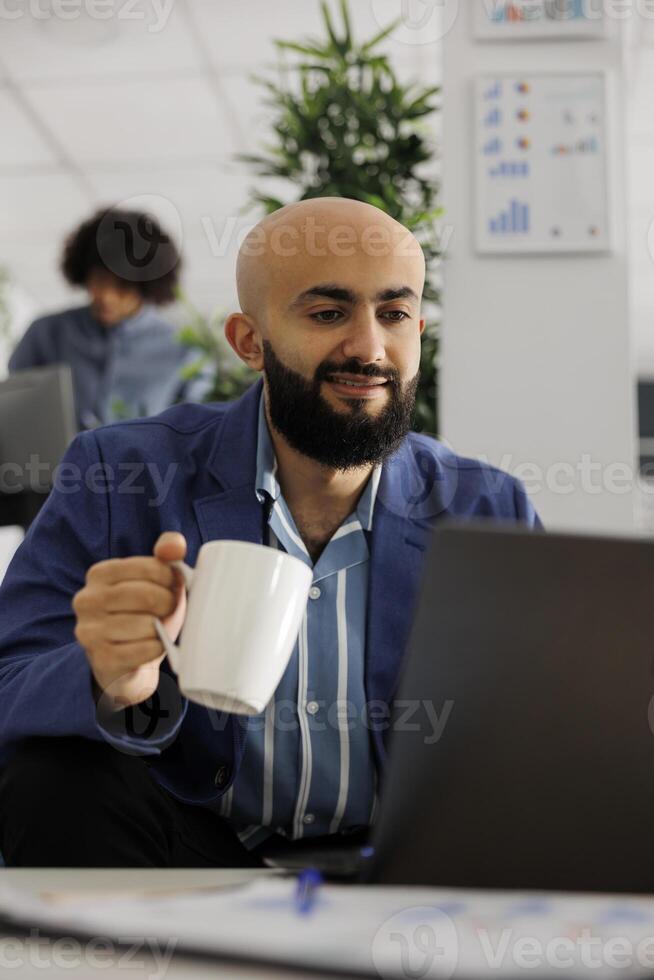 Smiling business company employee checking project financial report and drinking coffee. Arab start up entrepreneur holding tea mug and working at laptop in coworking space photo