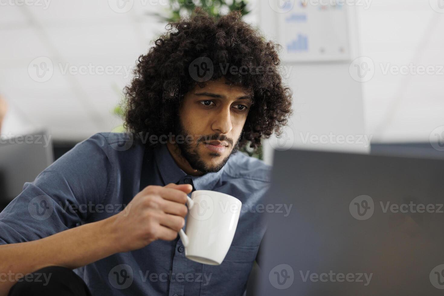 Entrepreneur drinking coffee while working on laptop to meet project deadline in business office. Arab start up company employee holding tea mug and researching data for analysis photo
