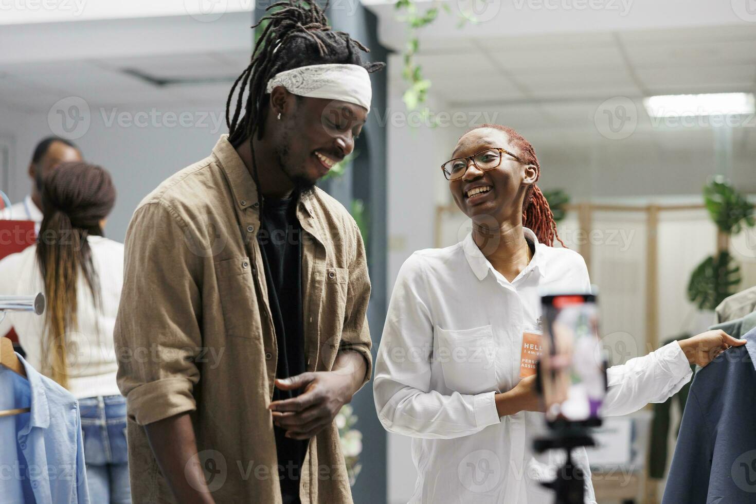 African american influencer and boutique owner live streaming on social media while showcasing outfits in showroom. Blogger and shop worker promoting fashion brand while recording video on smartphone photo