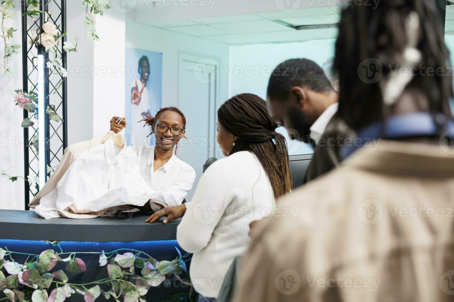 Clothing store african american customers waiting in line to pay for purchase at cash register. Mall employee working at checkout counter, holding apparel on hangers and chatting with clients photo