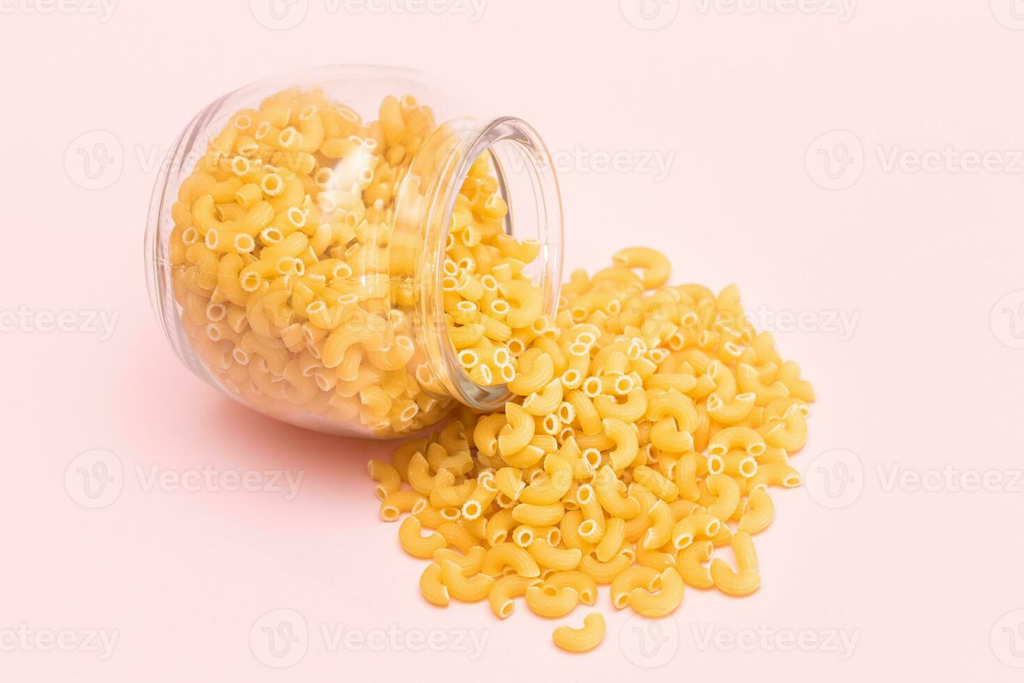 Uncooked Chifferi Rigati Pasta in Glass Jar on Pink Background. Fat and Unhealthy Food. Scattered Classic Dry Macaroni. Italian Culture and Cuisine. Raw Pasta photo