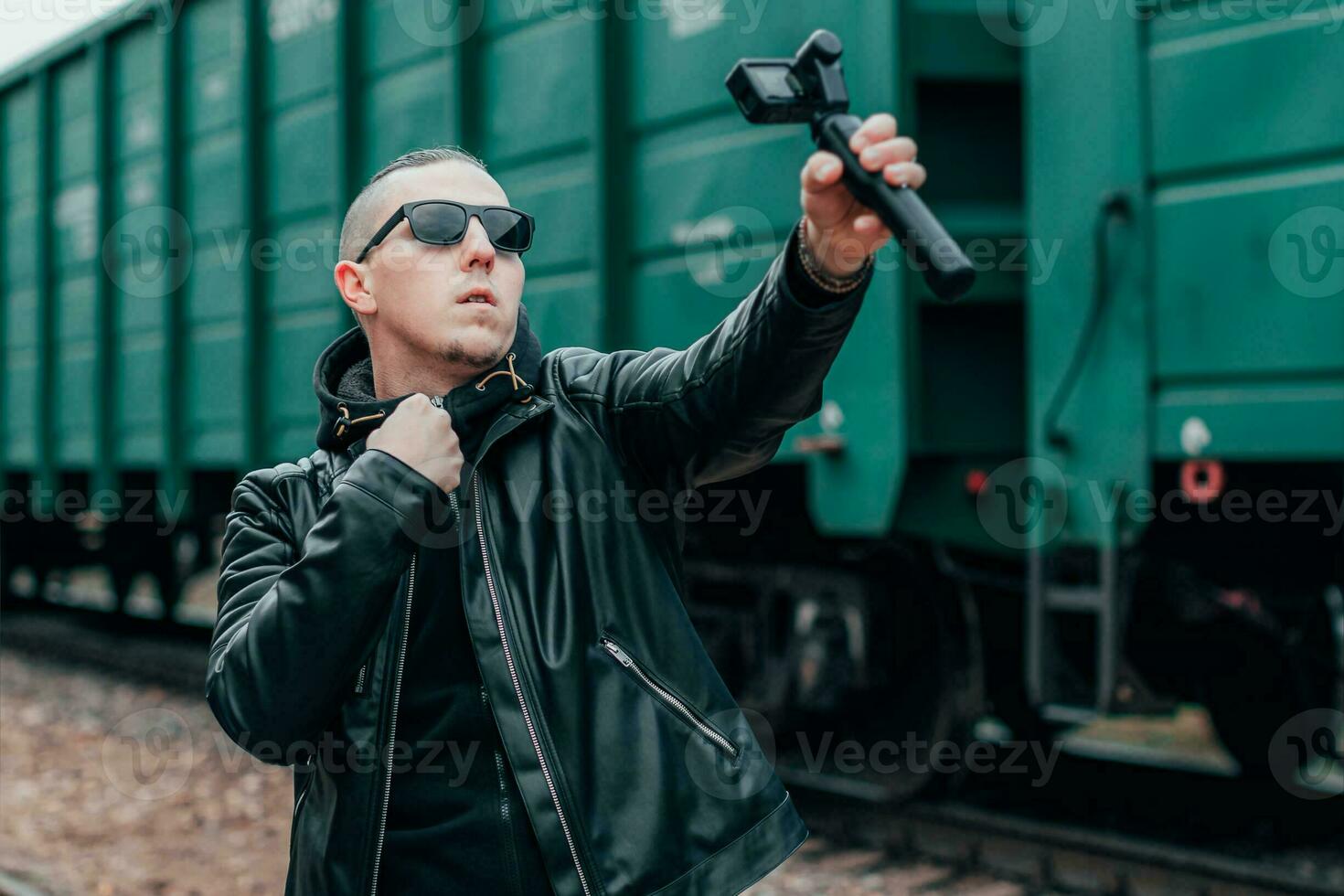 Handsome Guy in Black Clothes and Sunglasses Making Selfie or Streaming Video Using Action Camera with Gimbal Camera Stabilizer at Railway. Young Man Makes Photo Against the Cargo Wagons