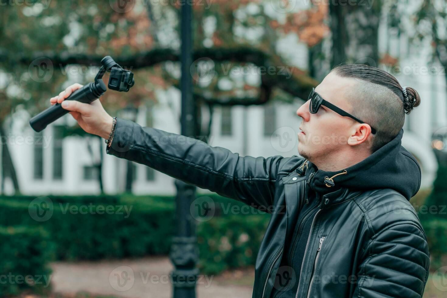 Blogger in Sunglasses Making Selfie or Streaming Video at the Autumn Park Using Action Camera with Gimbal Camera Stabilizer. Handsome Man in Black Clothes Making Photo