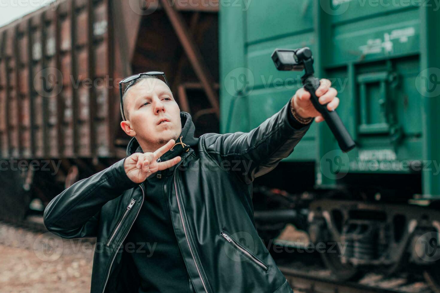 Handsome Guy in Black Clothes and Sunglasses Making Selfie or Streaming Video Using Action Camera with Gimbal Camera Stabilizer at Railway. Man Makes Photo Against the Cargo Wagons