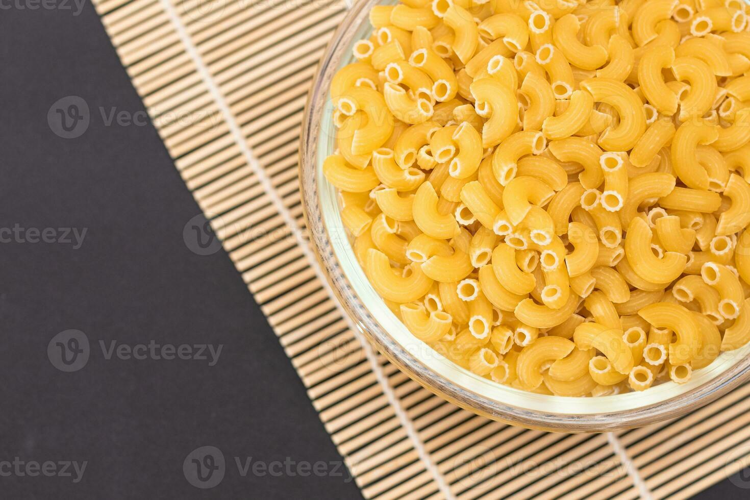 Uncooked Chifferi Rigati Pasta in Glass Jar on Bamboo Mat on Black Background. Fat and Unhealthy Food. Classic Dry Macaroni. Italian Culture and Cuisine. Raw Pasta photo