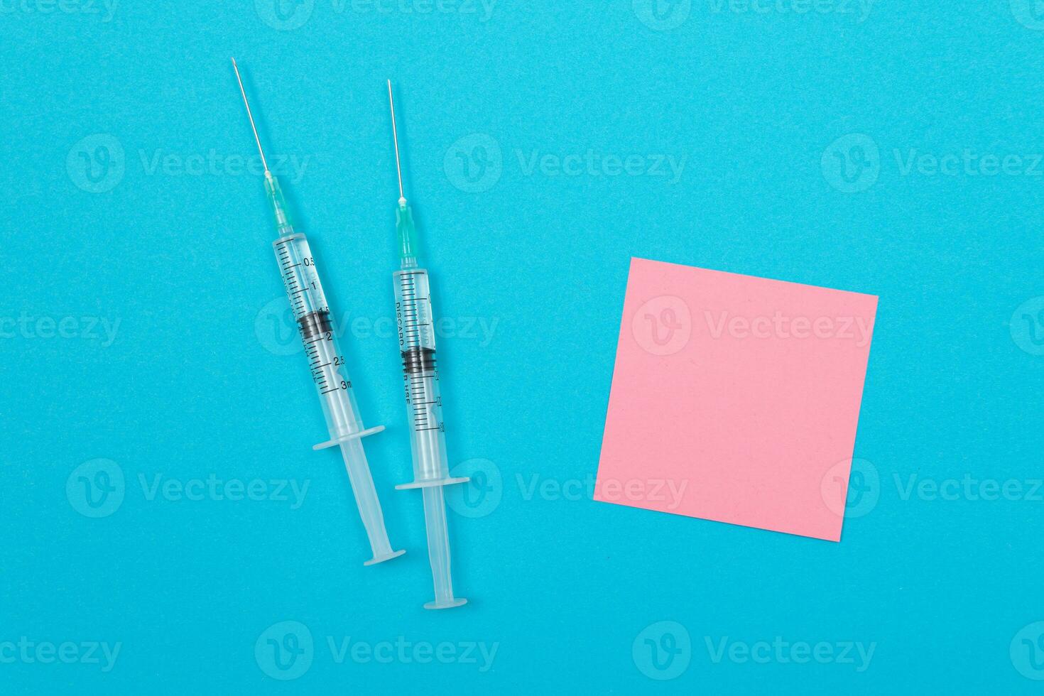 Vaccination, Immunology or Revaccination Concept Two Medical Disposable Syringe Lying on Blue Table in Doctor's Office in a Hospital or Clinic. Blank Pink Sticky Note Mock Up with Copy Space photo