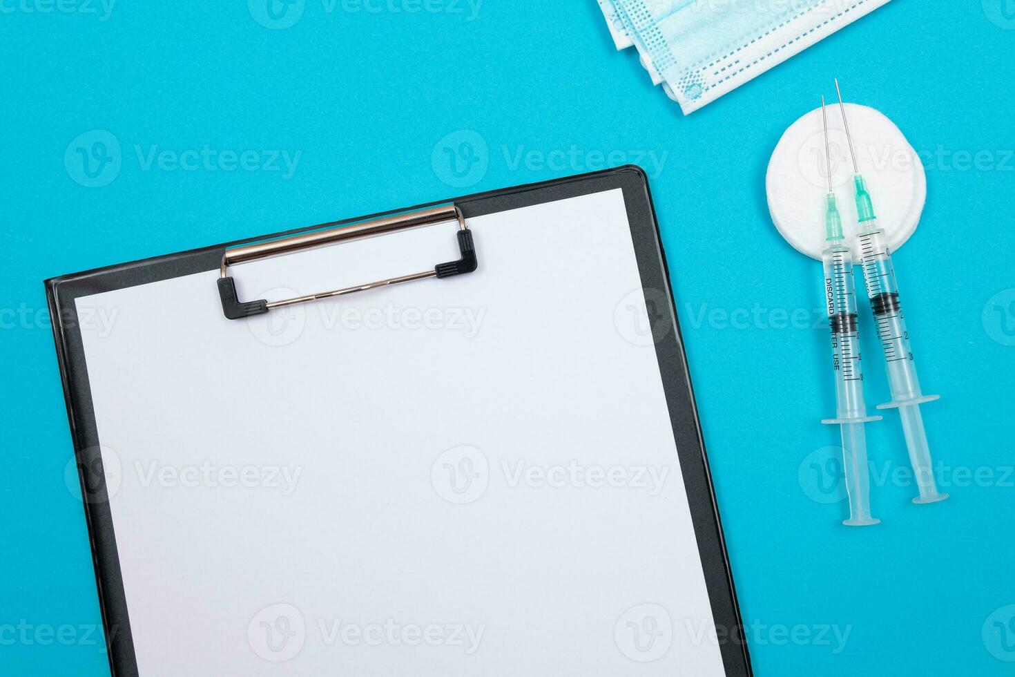 Vaccination, Immunology or Revaccination Concept Two Medical Syringe Lying on Blue Table in Doctor's Office in a Hospital or Clinic. Black Clipboard with Sheet of Paper Mock Up with Copy Space photo