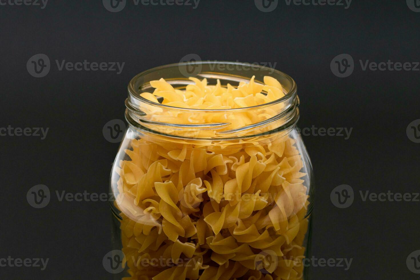 Uncooked Fusilli Pasta in Glass Jar on Black Background. Raw and Dry Macaroni. Unhealthy and Fat Food. Italian Culture photo