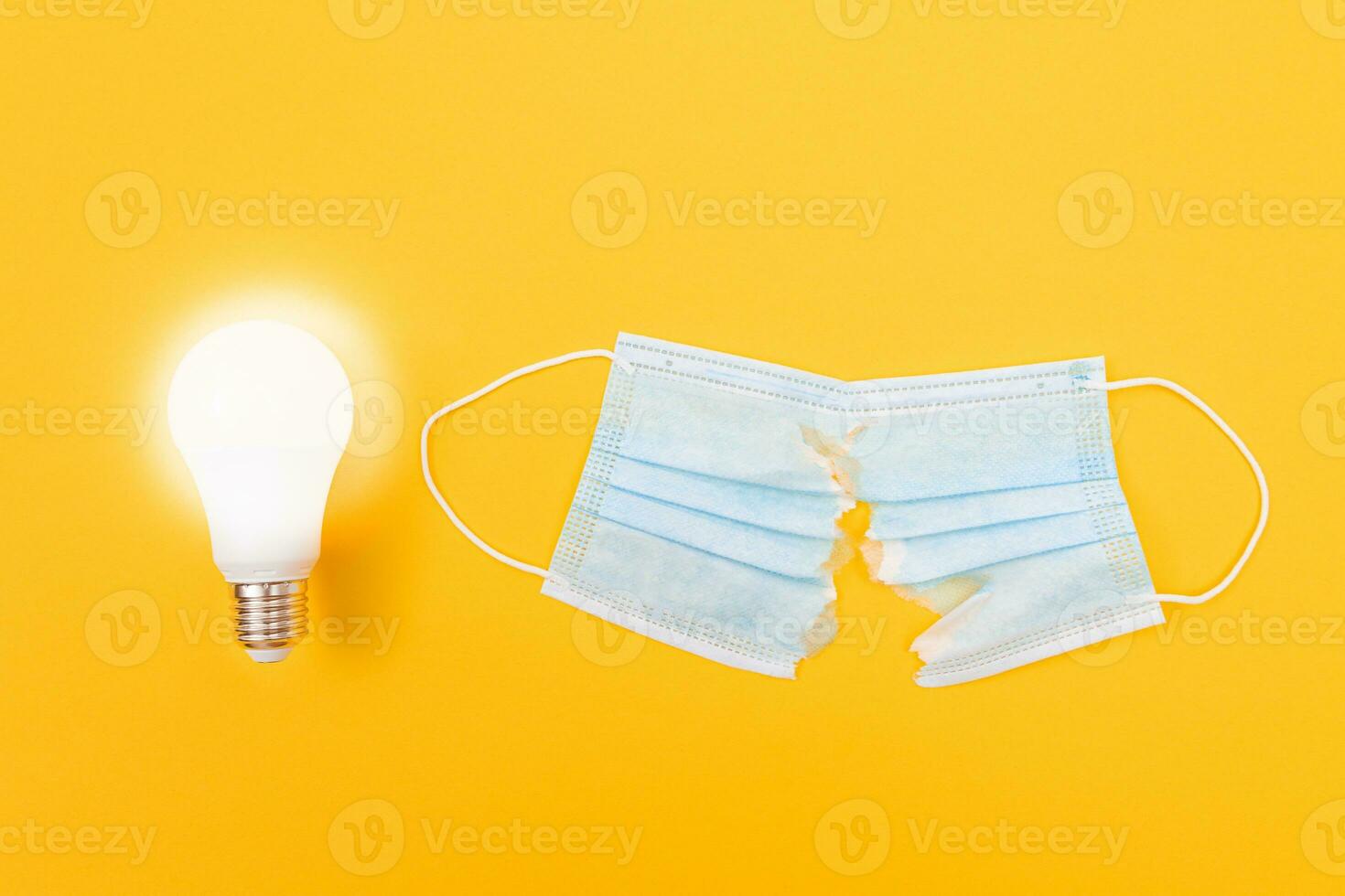 Glowing Power Saving Lamp with Torn Medical Face Mask Lying on Yellow Background Anti Mask Concept photo