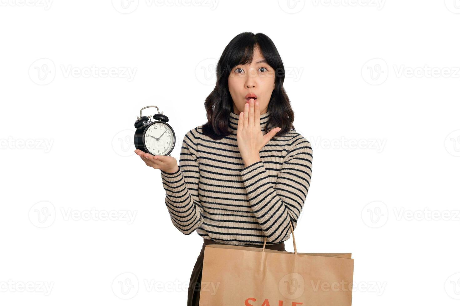 retrato de emocionado joven asiático mujer con suéter camisa participación alarma reloj y compras papel bolso aislado en blanco antecedentes. último ofertas conmocionado precio compras concepto foto