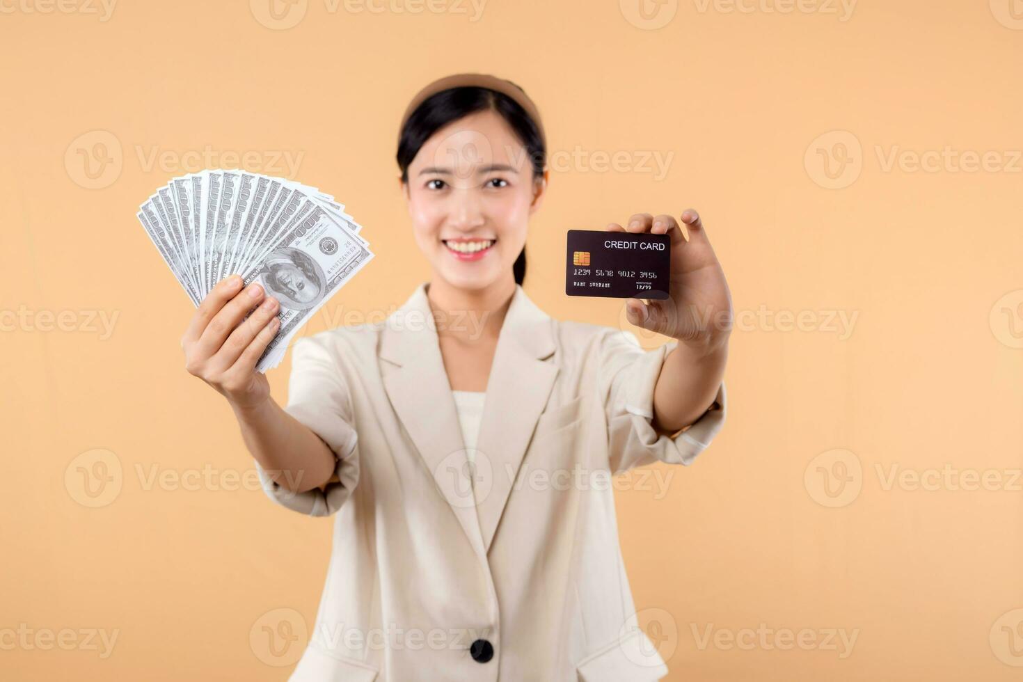 portrait of happy successful confident young asian business woman wearing white jacket holding cash money dollars and credit card standing over beige background. millionaire business, shopping concept photo