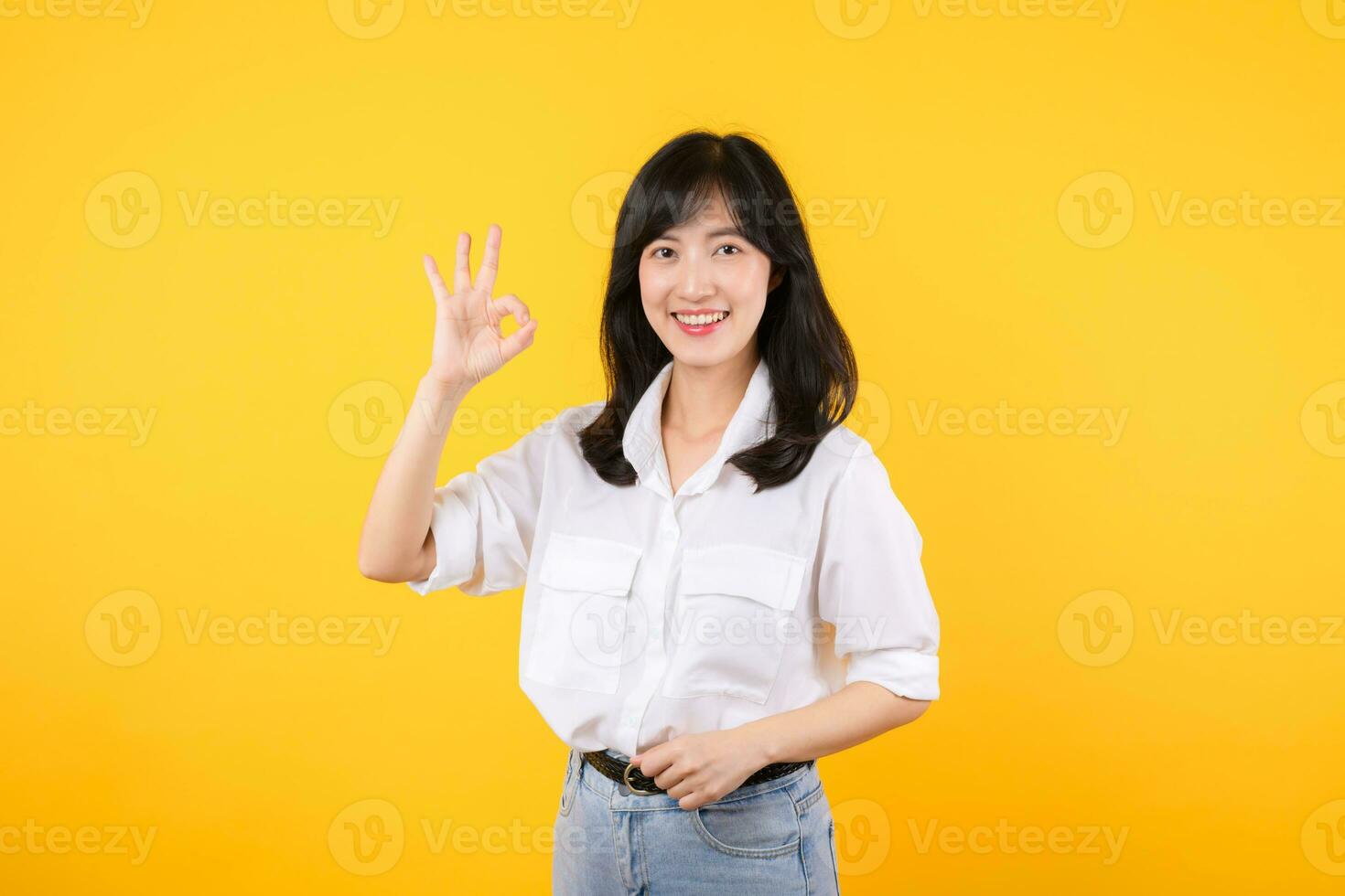 Young businesswoman hand sign okay gesture. Asian woman happy smile wearing white shirt and jean denim plants showing okay sign hand gesture isolated on yellow background. photo