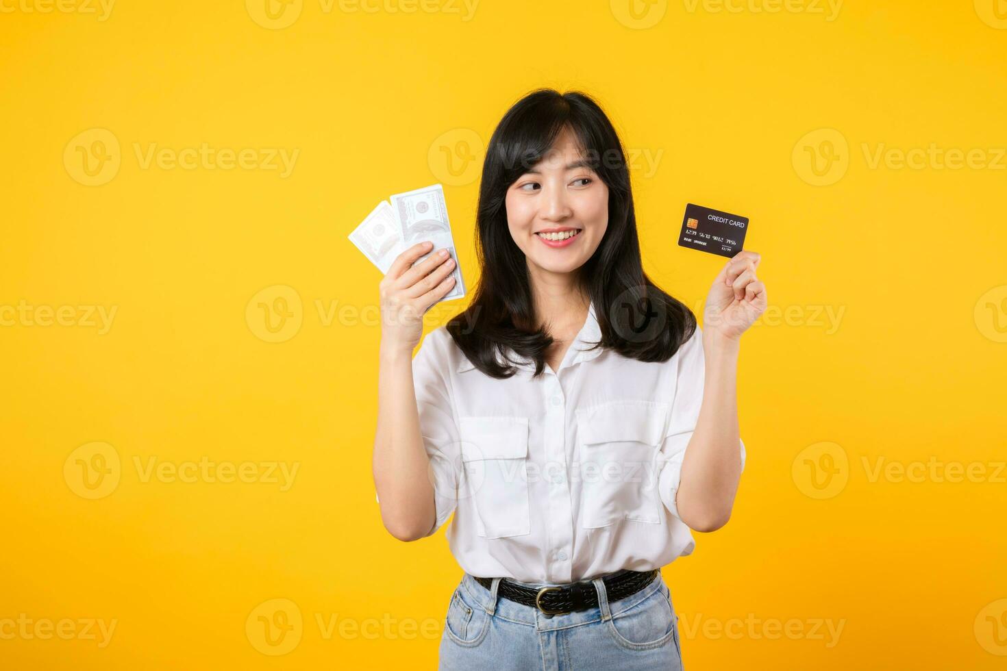 happy successful confident young asian woman happy smile wearing white shirt and denim jean holding cash money and credit card standing over yellow background. millionaire business, shopping concept. photo