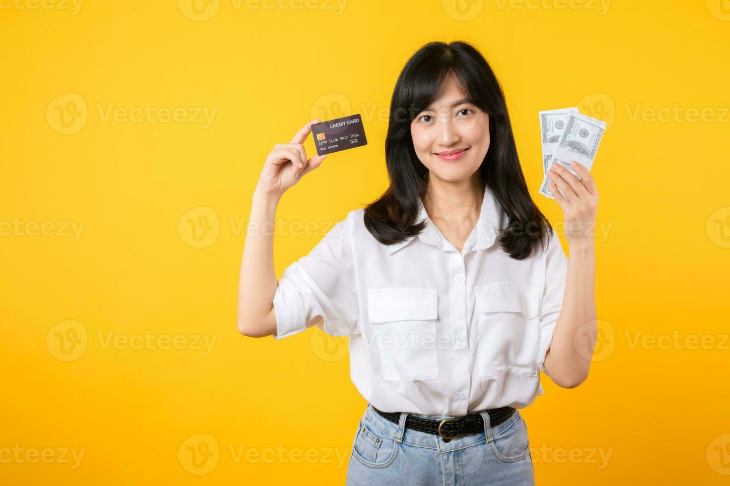 happy successful confident young asian woman happy smile wearing white shirt and denim jean holding cash money and credit card standing over yellow background. millionaire business, shopping concept. photo