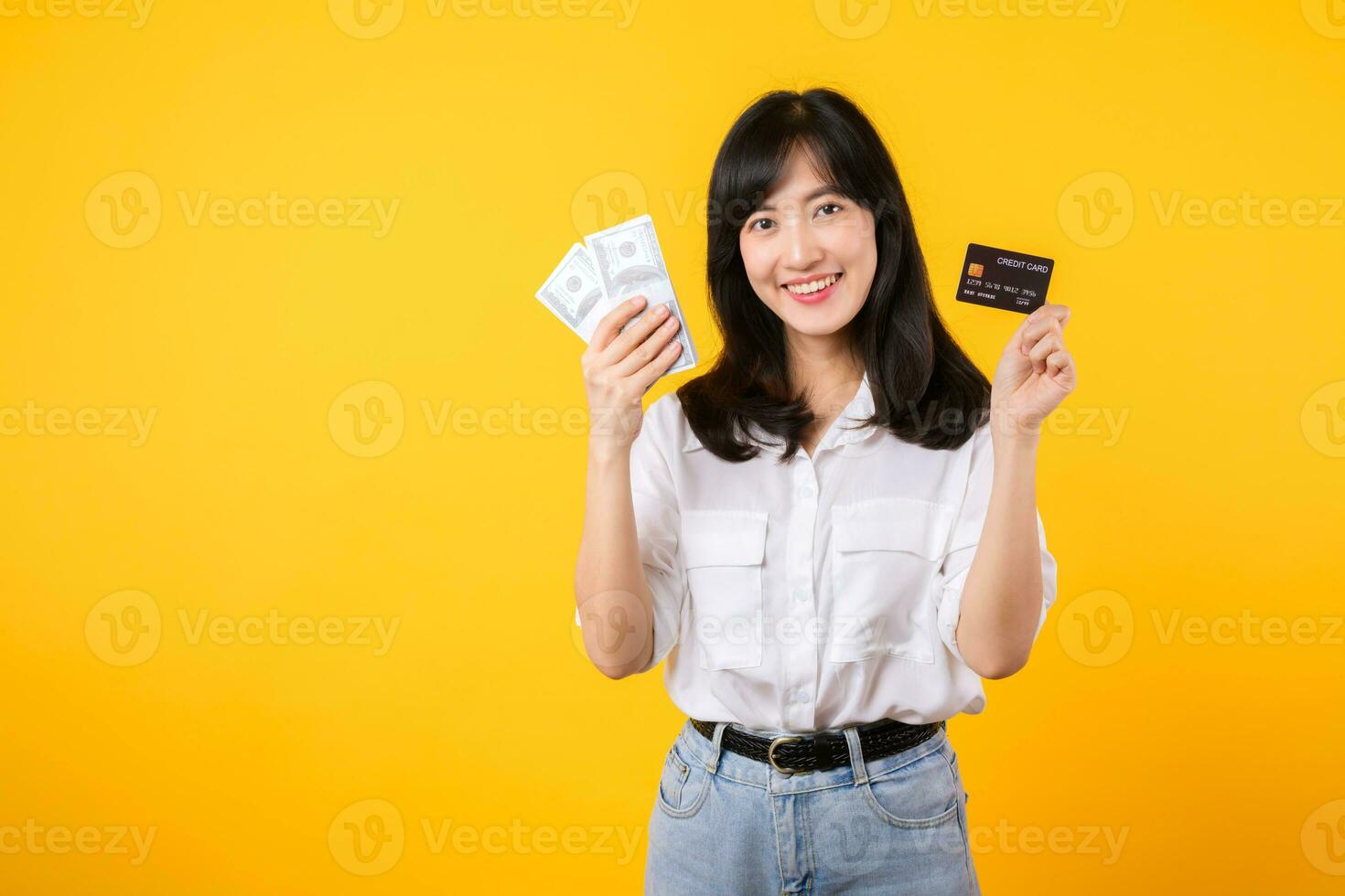 happy successful confident young asian woman happy smile wearing white shirt and denim jean holding cash money and credit card standing over yellow background. millionaire business, shopping concept. photo