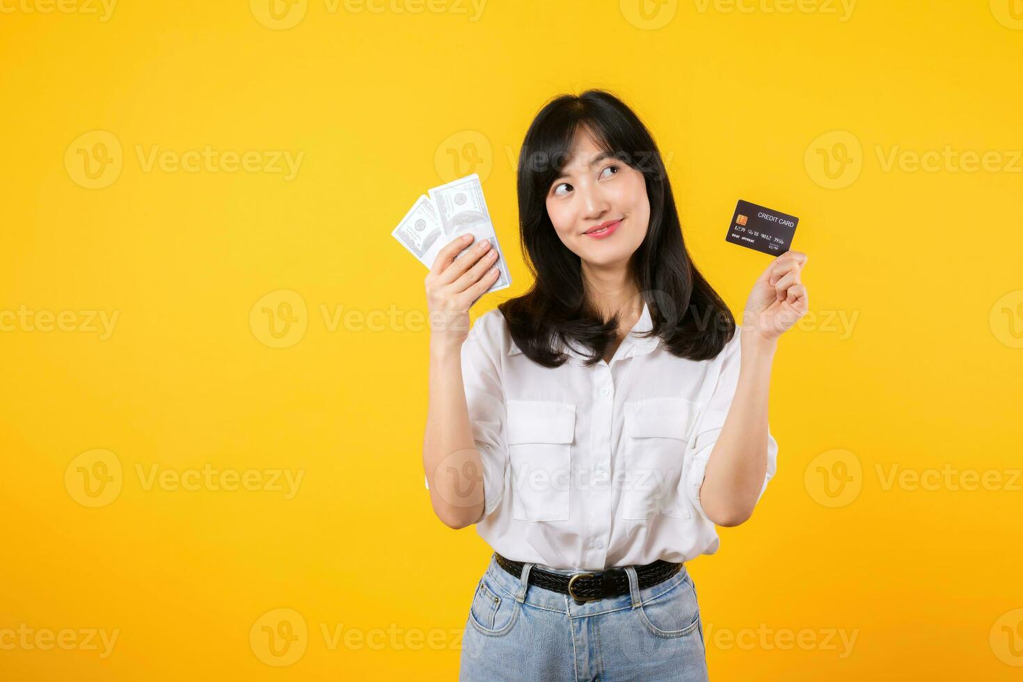 happy successful confident young asian woman happy smile wearing white shirt and denim jean holding cash money and credit card standing over yellow background. millionaire business, shopping concept. photo