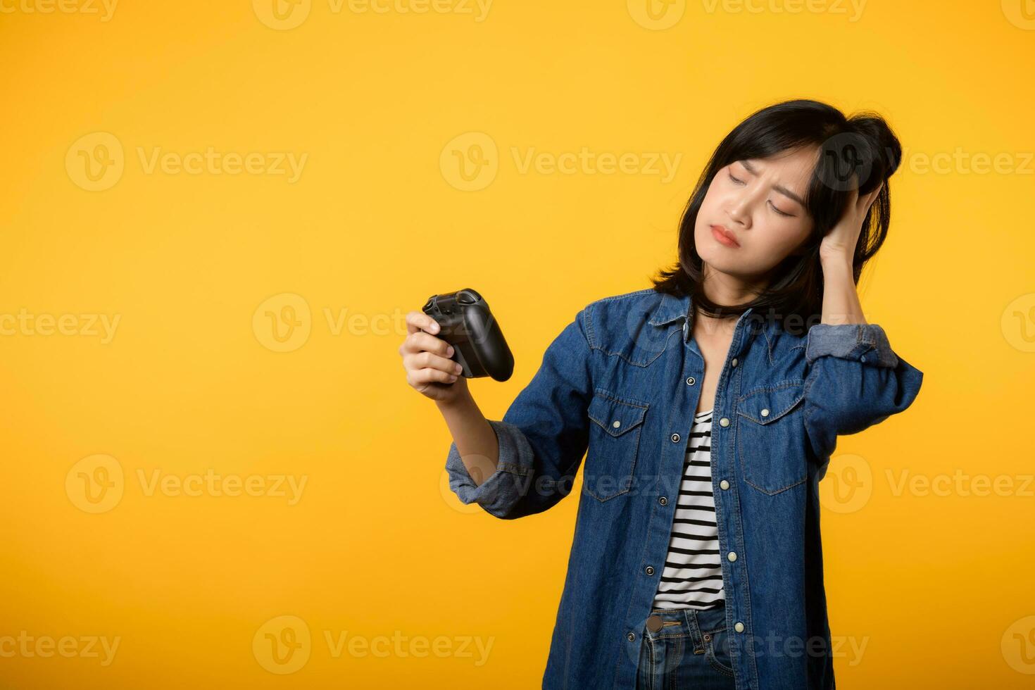 Portrait young asian woman with doubt face wearing denim clothes holding joystick controller and playing video game. Fun and relax hobby entertainment lifestyle concept. photo