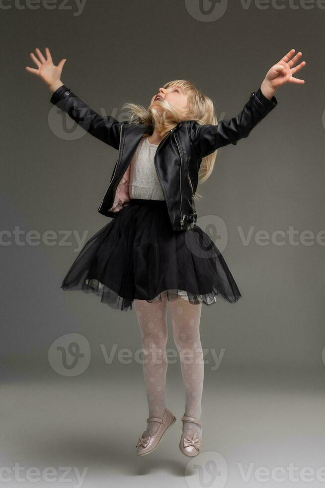 Little blonde girl in a white dress and black leather jacket is posing standing over a gray background. photo