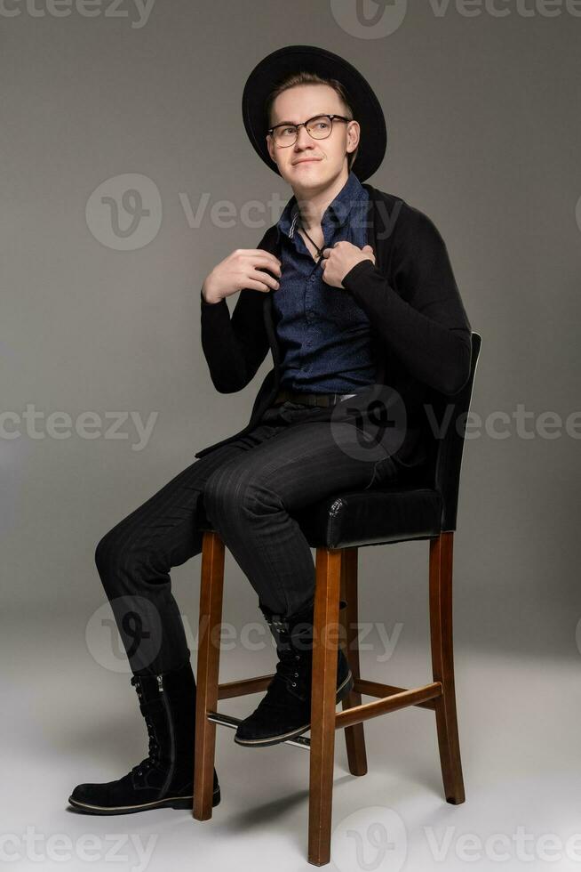 Brunet man in a casual dark clothes posing over a grey background sitting on the bar chair. photo