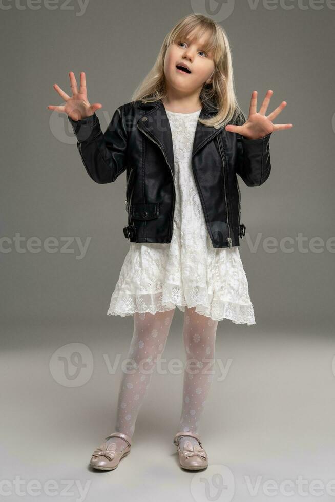 Little blonde girl in a white dress and black leather jacket is posing standing over a gray background. photo