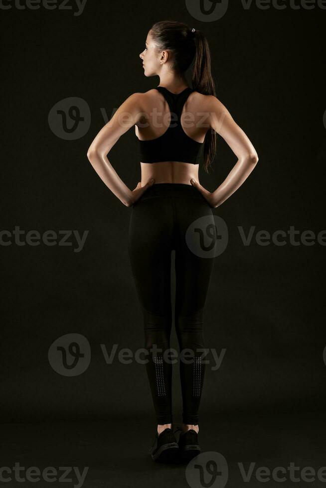 Brunette woman in black leggings, top and sneakers is posing against a black background. Fitness, gym, healthy lifestyle concept. Full length. photo